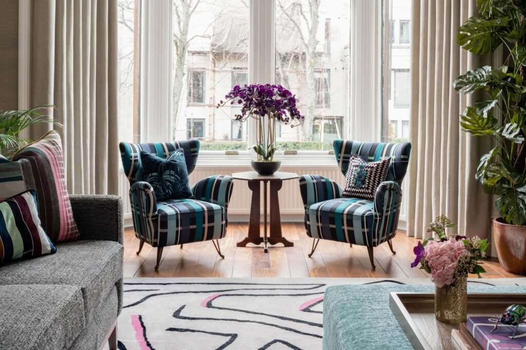 Bright, eclectic living room with two teal plaid armchairs, a purple orchid, grey sofa, abstract rug, and large windows.