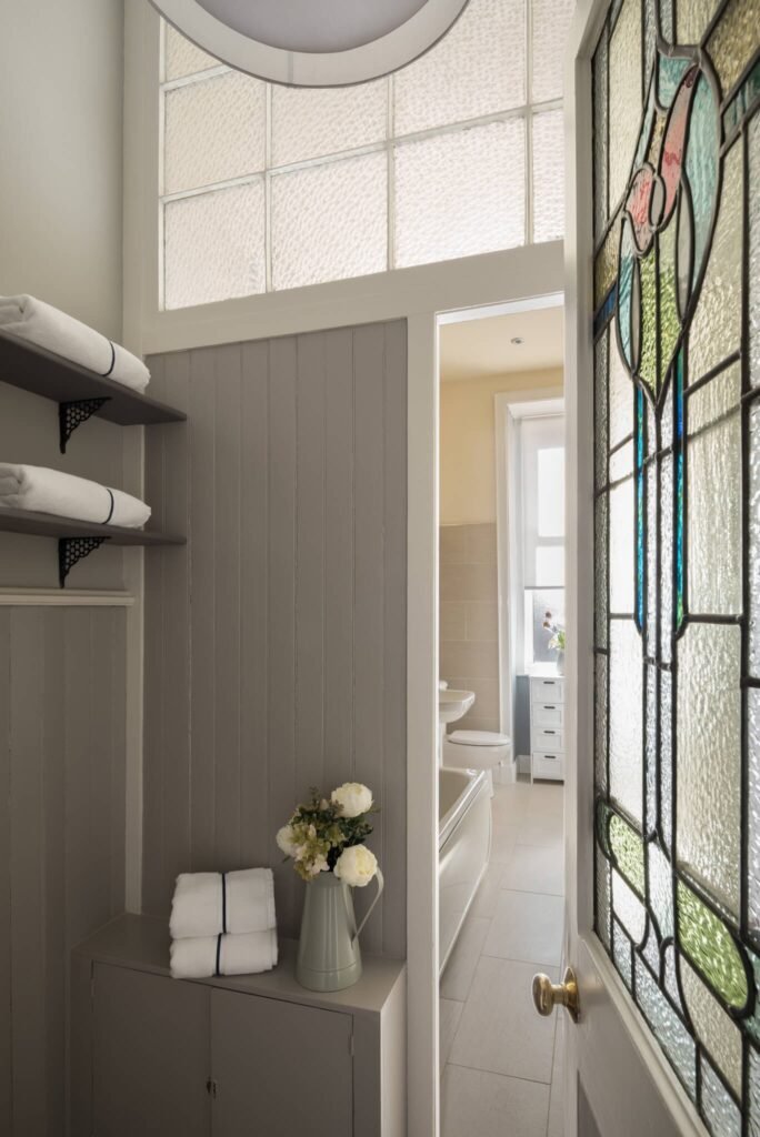 Cozy grey bathroom entryway with stained glass door, textured glass window, stacked white towels, and fresh flowers.