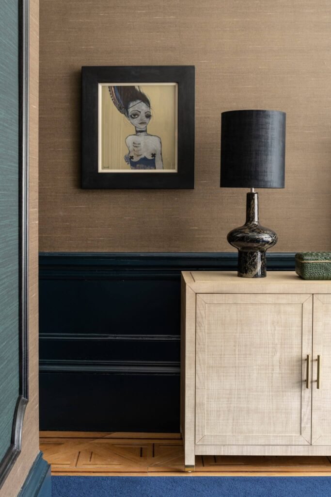 Eclectic room with brown textured wallpaper, dark blue wainscoting, framed art, a patterned black lamp, and light wood cabinet.