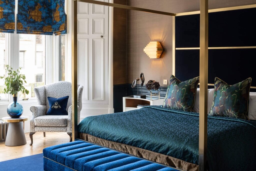 Elegant bedroom with a gold four-poster bed, deep teal bedding, blue velvet bench, and detailed window blinds.