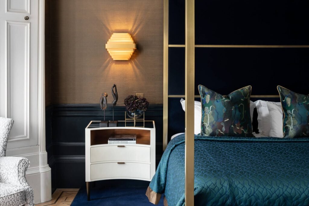 Sophisticated bedroom with brass four-poster bed, dark blue headboard, curved nightstand, abstract sculptures, and a geometric wall sconce.