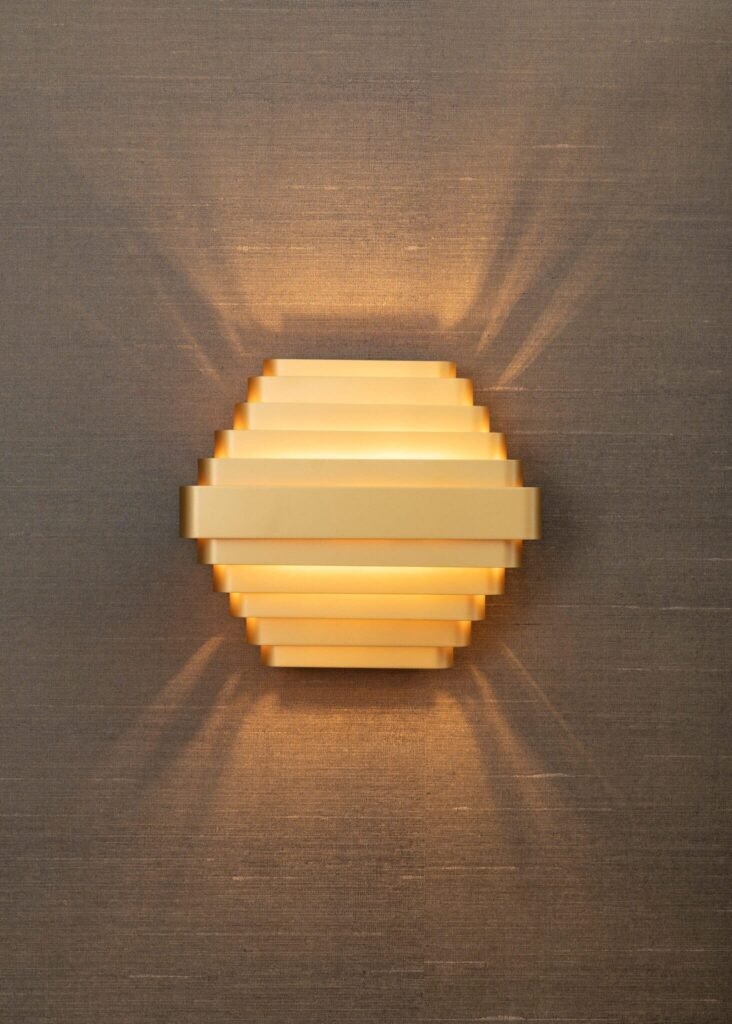 Brass, tiered wall sconce casting a warm, fanned light pattern on a textured brown fabric wall covering.