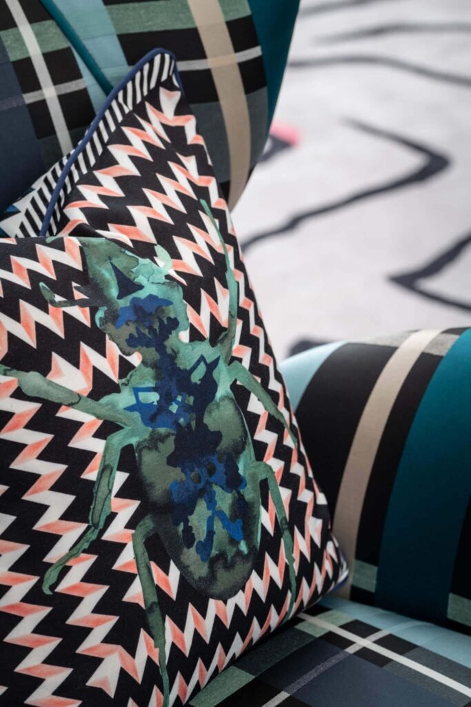 Vibrant pillow with teal spider and black-white-coral zig-zag pattern, resting on a plaid armchair.
