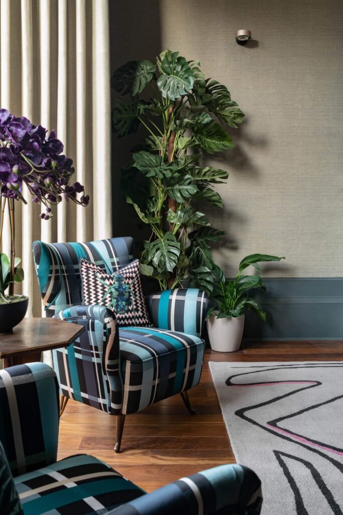 Vibrant blue and black plaid armchairs, large Monstera plant, purple orchids, and abstract rug in a modern room.
