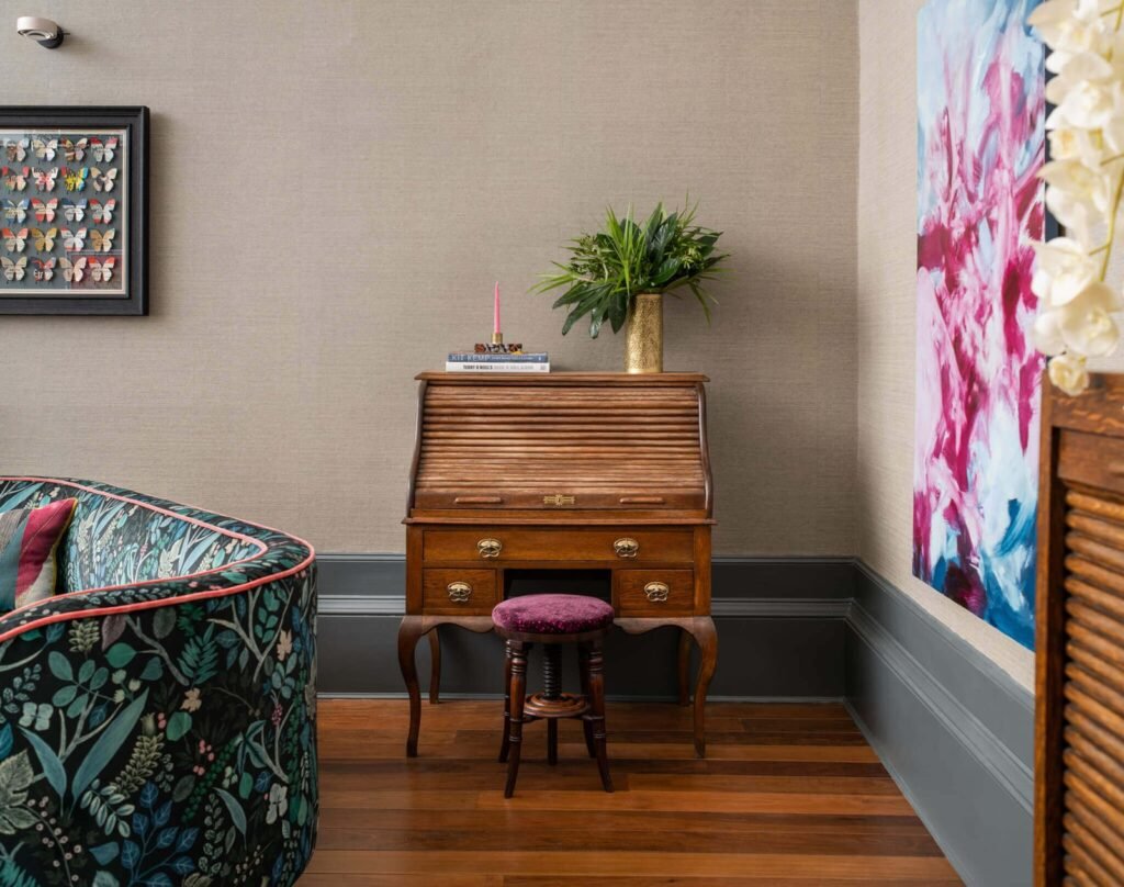Interior with a vintage roll-top desk, purple velvet stool, botanical sofa, framed butterfly art, and abstract painting.