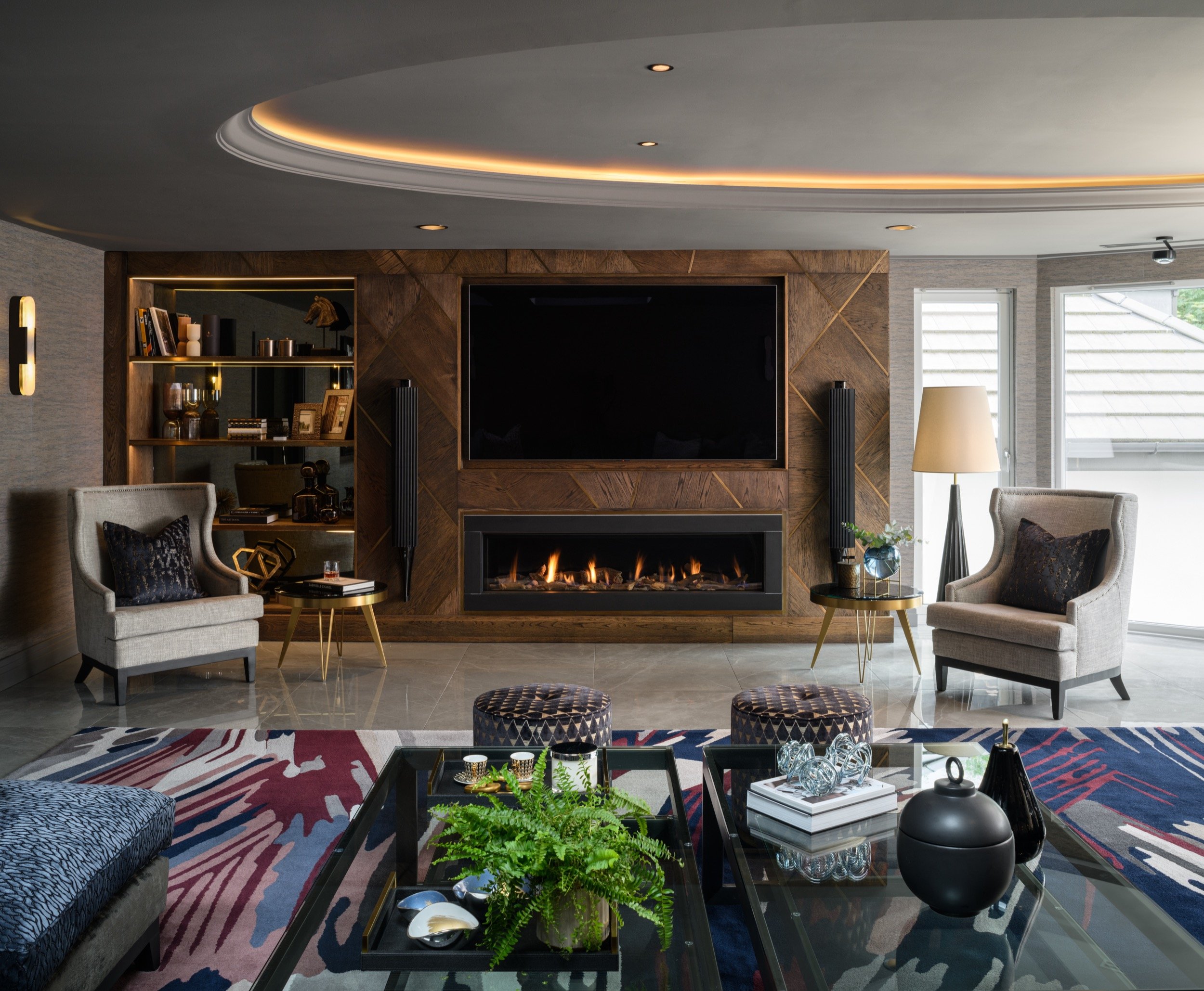 Elegant living room with a large TV and modern fireplace in a geometric wood wall, accompanied by chic armchairs and a patterned rug.