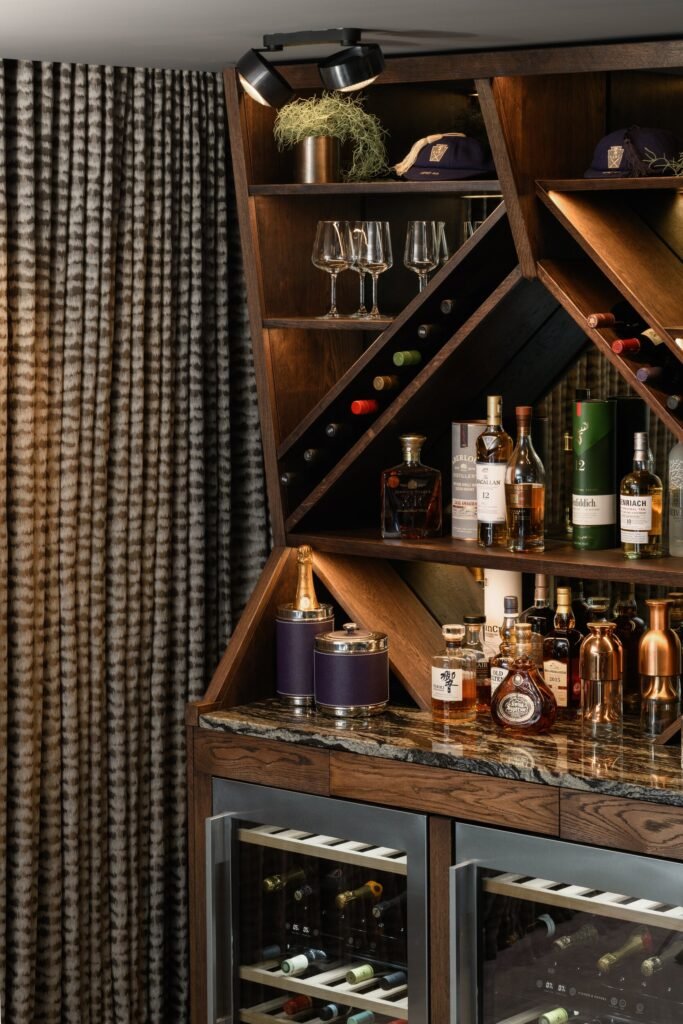 Modern dark wood bar with granite counter, illuminated shelves of whiskey & wine, built-in fridges, and patterned drapes.