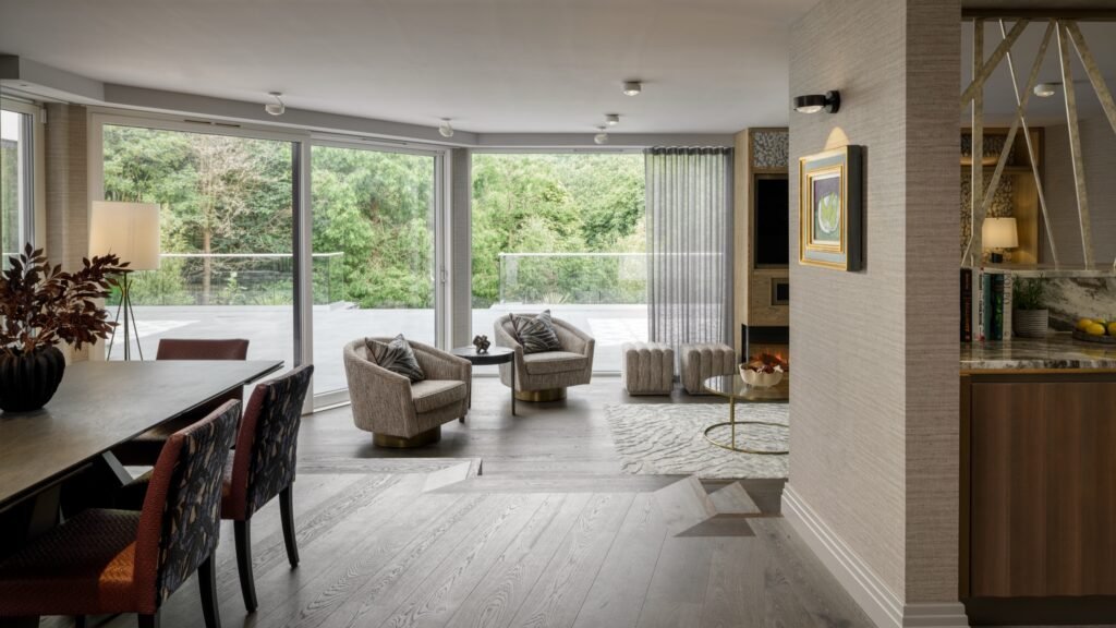 Contemporary open-plan living & dining room with large windows showcasing a lush forest view, modern furniture, and light wood flooring.