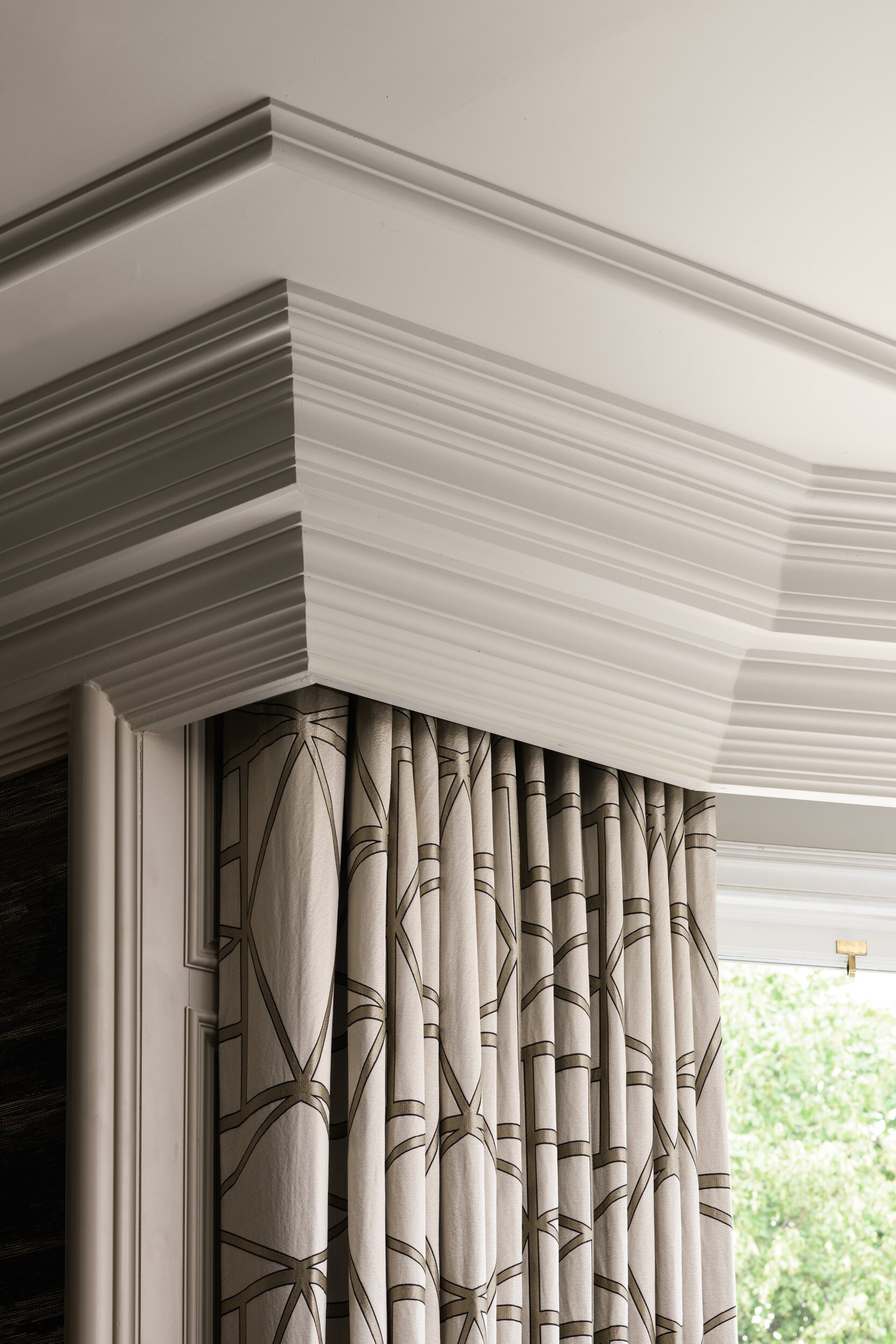 Layered white crown molding above geometric patterned curtains by a window, with a glimpse of green outside.