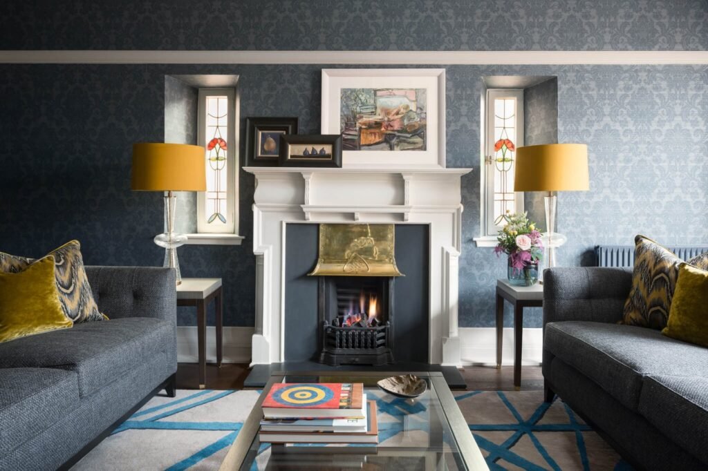 Elegant blue living room with damask wallpaper, a cozy fireplace, grey sofas, vibrant yellow lamps, and stained glass windows.