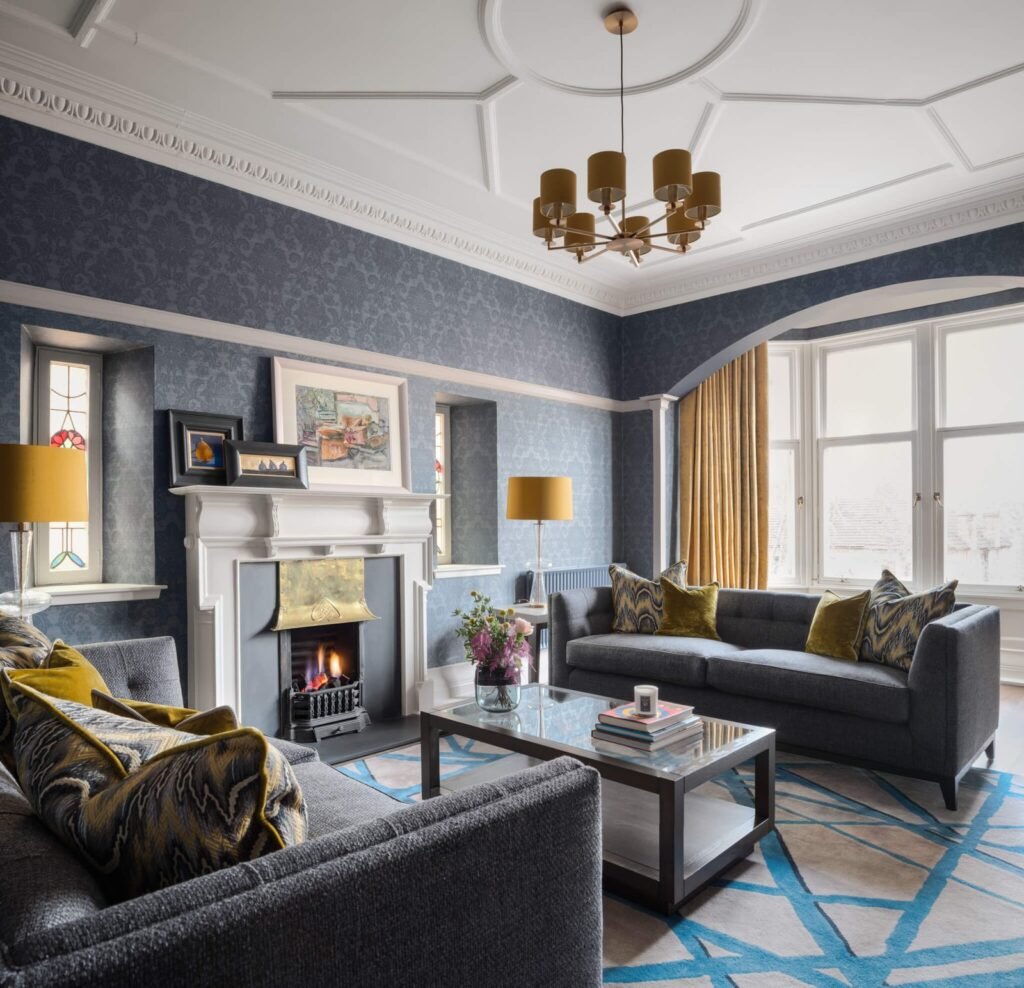 Elegant living room with blue patterned wallpaper, grey sofas, ornate ceiling, fireplace, gold accents, bay window, and abstract rug.
