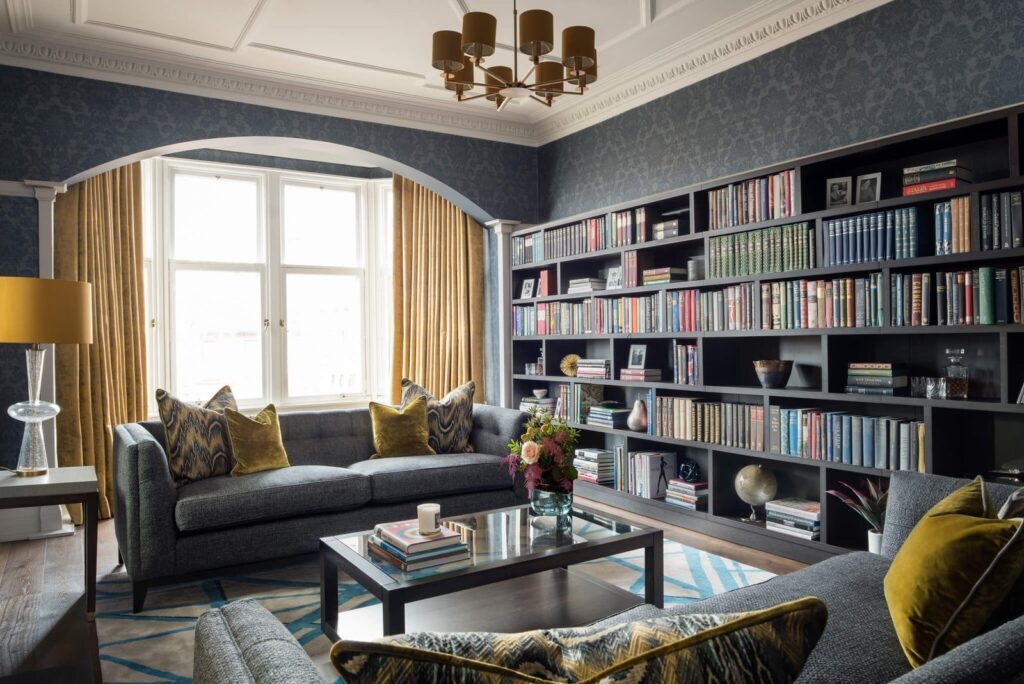 Sophisticated living room with full-wall bookshelf, grey sofa, gold accents, dark blue wallpaper, and bay window.