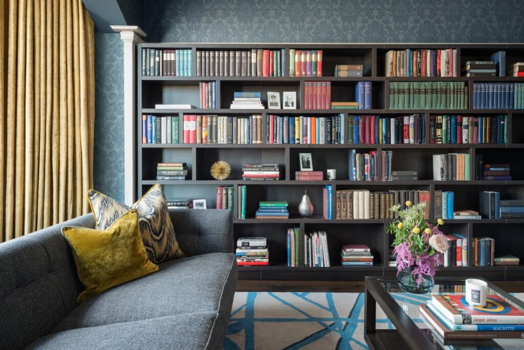 Sophisticated living room with a wall of dark bookshelves filled with books, grey sofa, yellow velvet pillows, and gold curtains.