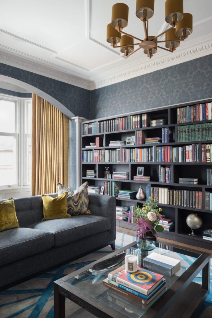 Elegant living room with dark blue damask wallpaper, a grey sofa, custom bookshelves, gold curtains, and a glass coffee table.