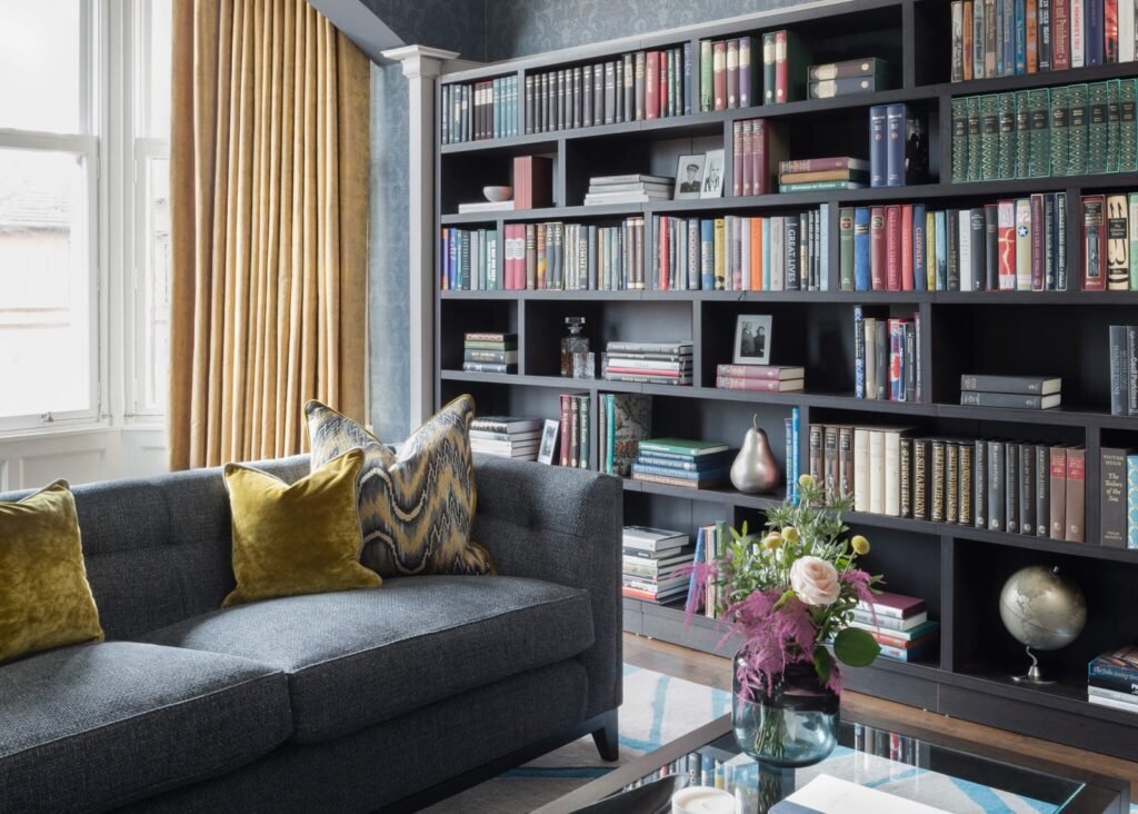 Cozy living room with dark gray sofa, gold cushions, gold curtains, and a dark bookshelf packed with books and decor.