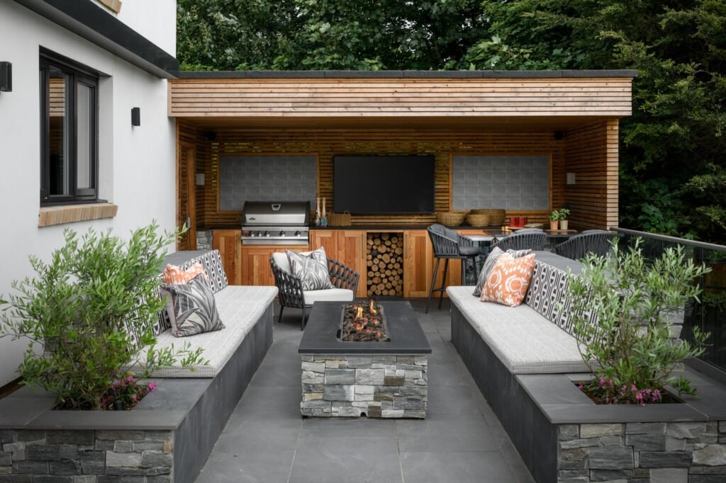 Luxurious outdoor living area featuring a covered kitchen with grill, built-in benches, stone fire pit, and integrated planters.