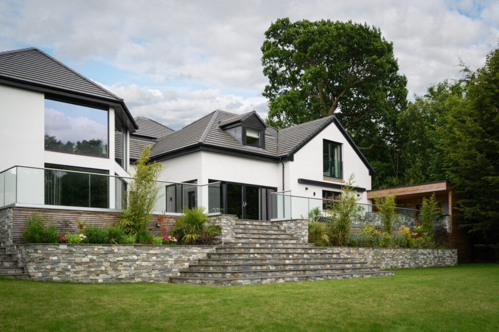 Modern white house exterior with large windows, glass balcony, tiered stone garden, and green lawn.