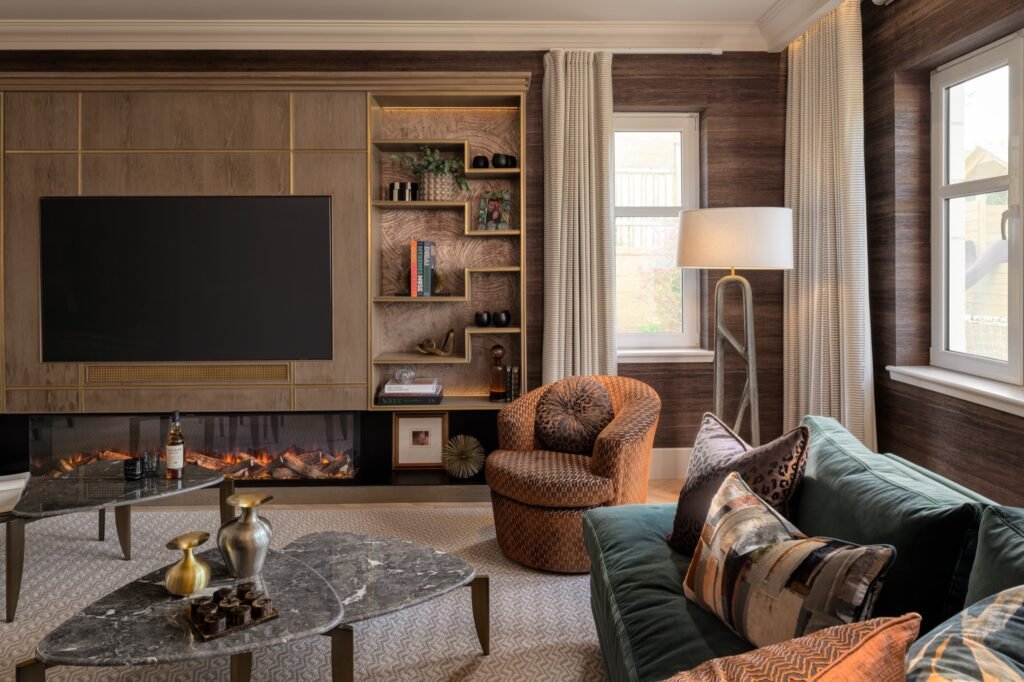 Elegant living room with wood media wall, TV, fireplace, green velvet sofa, patterned armchair, and marble coffee tables.