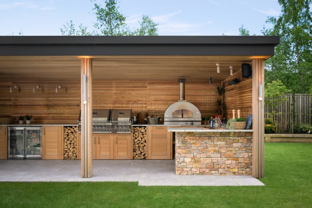 Covered outdoor kitchen with wood-paneled walls, pizza oven, grills, drinks fridge, stone island, and log storage.
