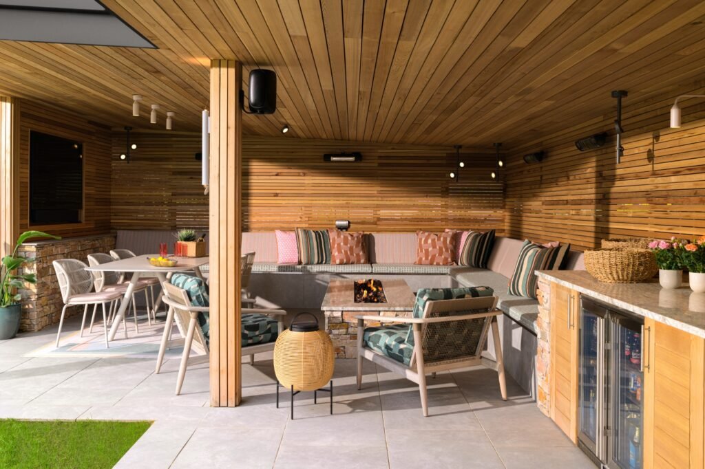 Modern covered outdoor living area with wood paneling, built-in L-shaped sofa, stone fire pit, dining table, and bar.
