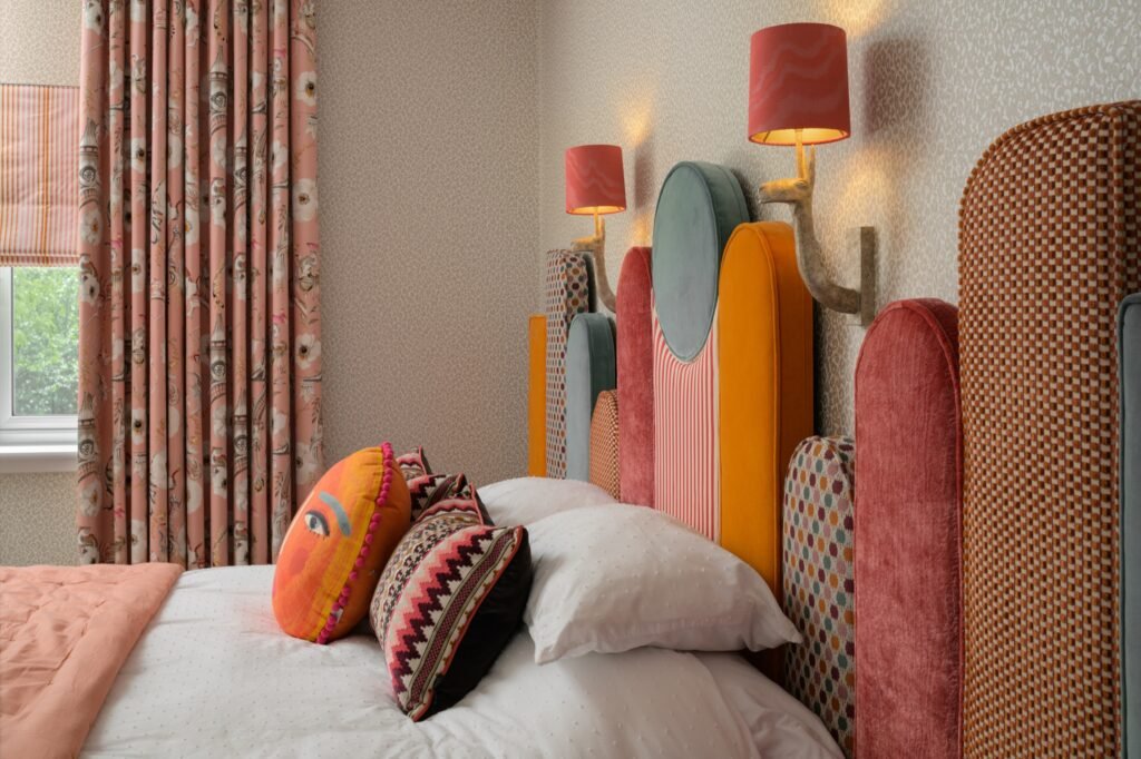 Eclectic bedroom: colorful segmented headboard, pink floral curtains, patterned wallpaper, and animal-head wall lamps.
