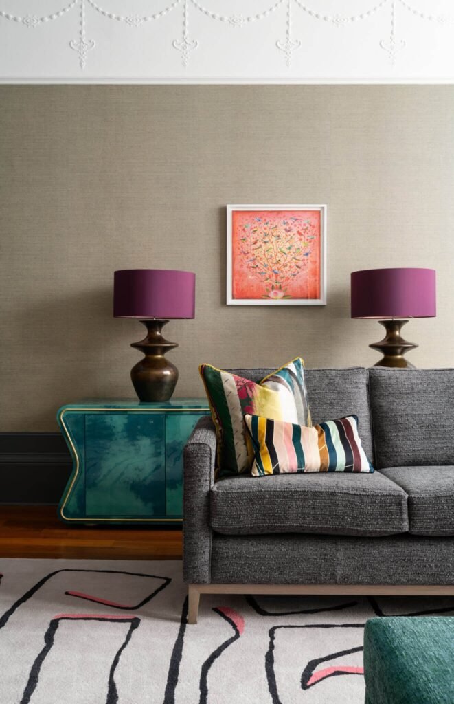 Modern eclectic living room: grey sofa, vibrant pillows, teal cabinet, purple lamps, colorful bird artwork, textured wallpaper.
