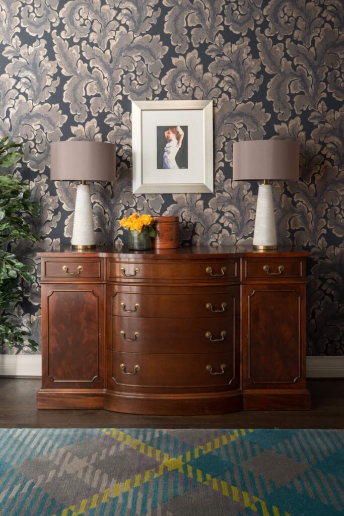 Antique wood sideboard with two lamps, framed art, and flowers against metallic damask wallpaper. A vibrant geometric rug adds color.