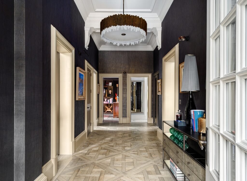 Elegant, long hallway with dark textured walls, light geometric wood floor, an ornate bronze chandelier, and open doorways.