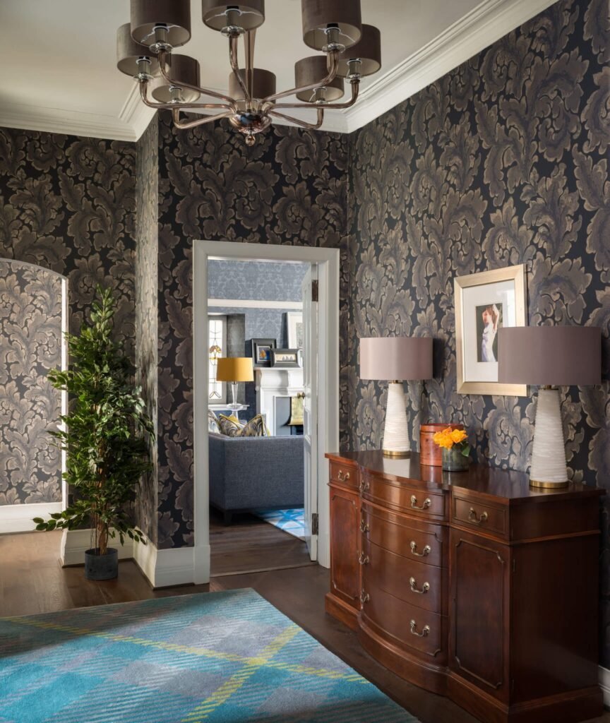 Elegant dark entryway with damask wallpaper, antique wooden sideboard, multi-arm chandelier, and blue plaid rug leading to living room.