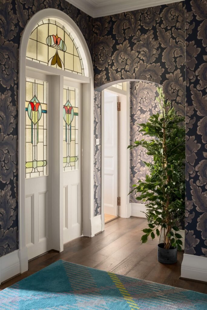 Elegant hallway with Art Nouveau stained glass entry, dark floral wallpaper, potted plant, wood floor, teal rug.
