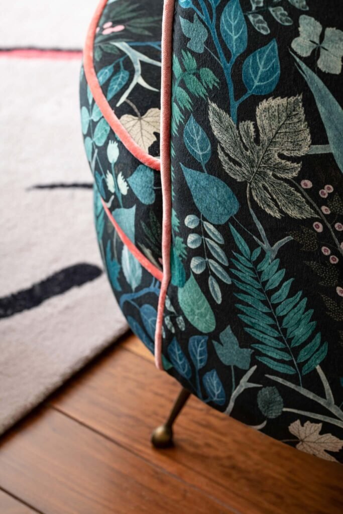 Close-up of an upholstered furniture piece with vibrant dark botanical fabric, teal/green leaves, and coral piping on a wood floor.