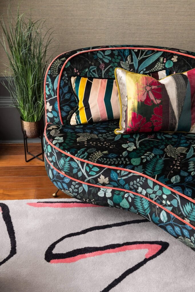Dark botanical sofa with bright coral piping, vibrant cushions, tall plant in copper pot, and abstract grey rug.