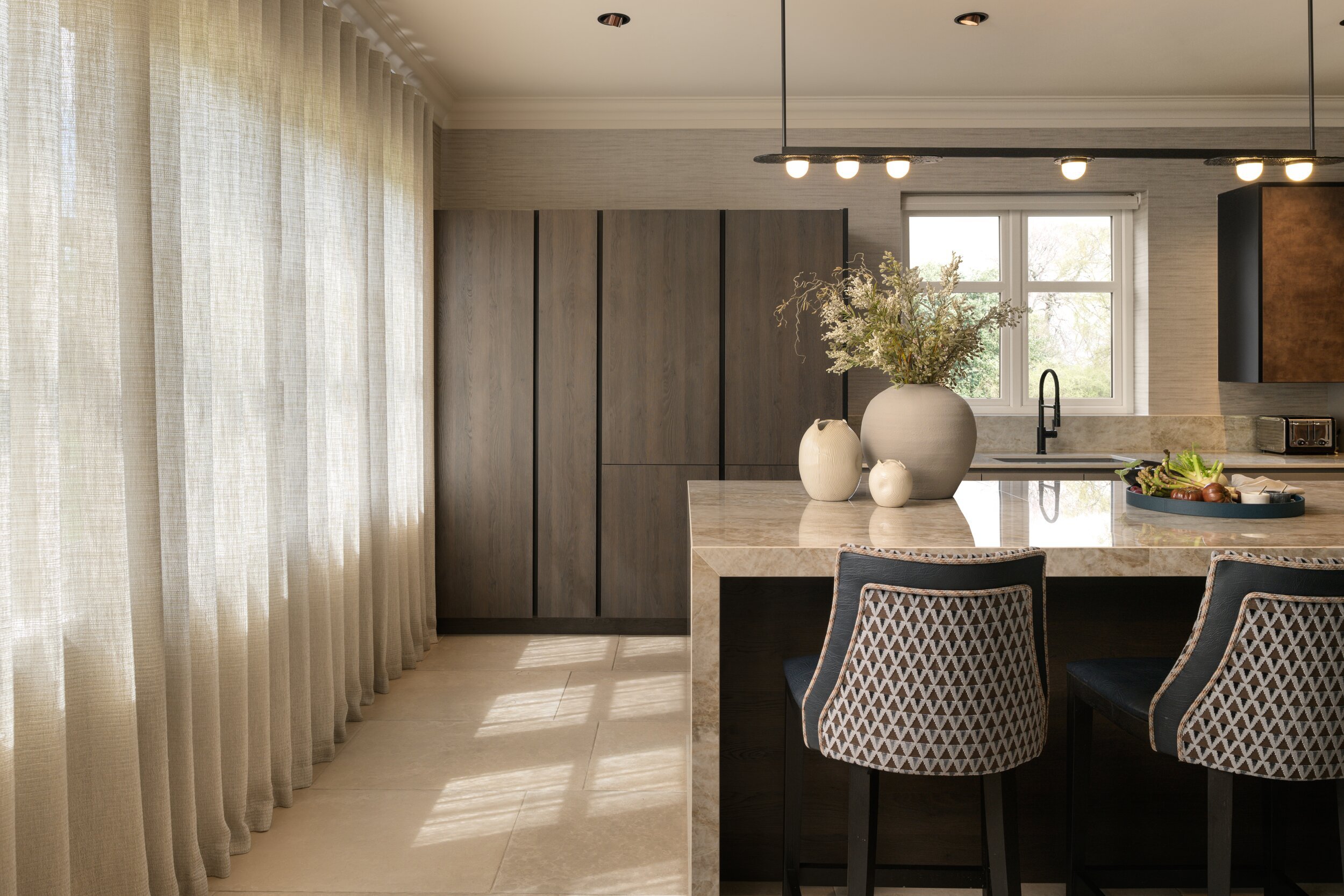 Stylish modern kitchen with marble island, dark wood cabinetry, patterned bar stools, and sheer curtains.
