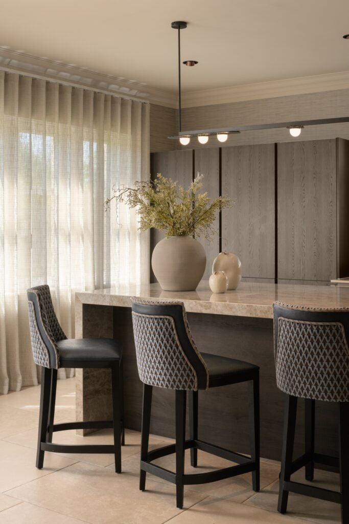 Stylish modern kitchen with marble island, dark wood cabinetry, patterned bar stools, and sheer curtains.
