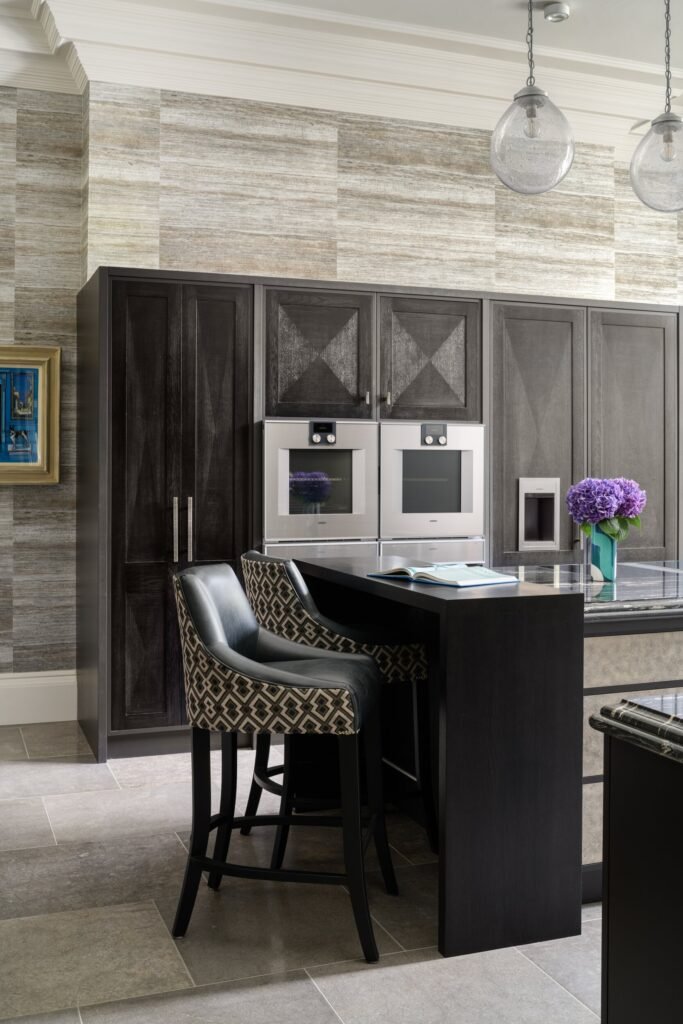 Sleek modern kitchen with dark cabinets, dual built-in ovens, wood-look walls, and an island with patterned bar stools.