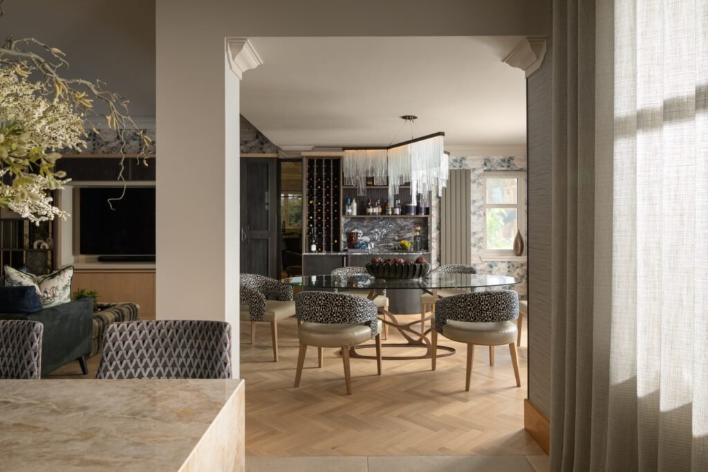 Luxury open-plan dining & living room with glass table, leopard chairs, modern chandelier, built-in bar, herringbone floor.
