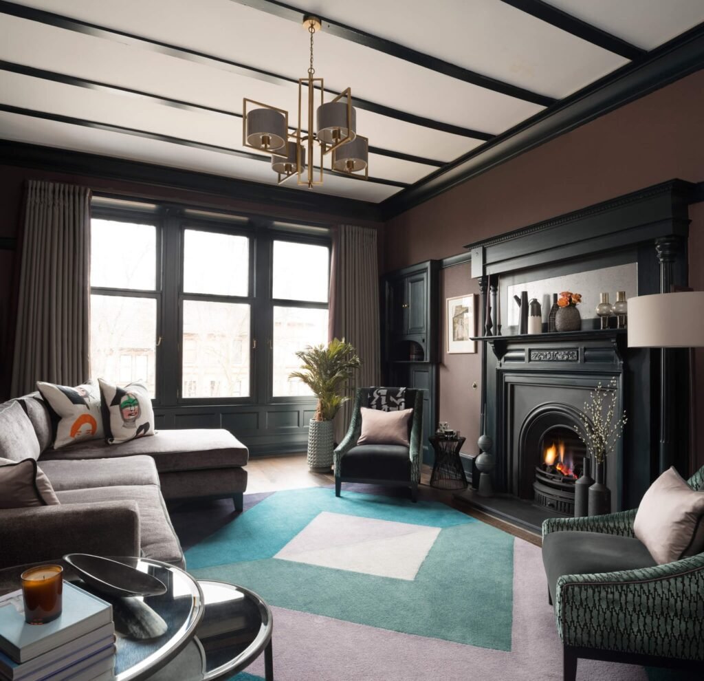 Chic, moody living room with deep brown walls, cozy fireplace, grey sectional sofa, vibrant geometric rug, and dark beamed ceiling.
