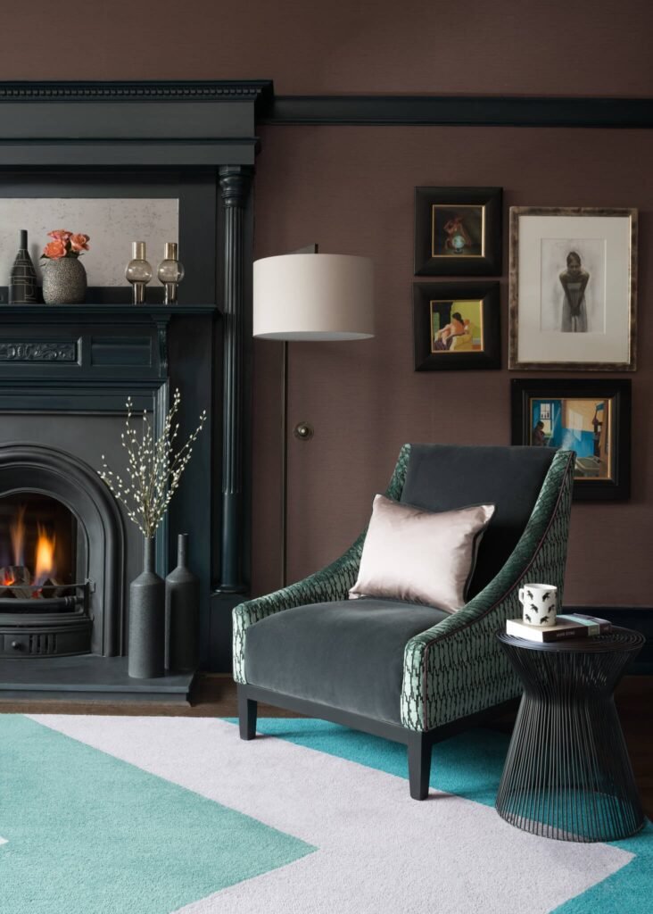 Luxurious living room with dark teal velvet armchair, burning fireplace, brown walls, gallery art, and geometric teal rug.