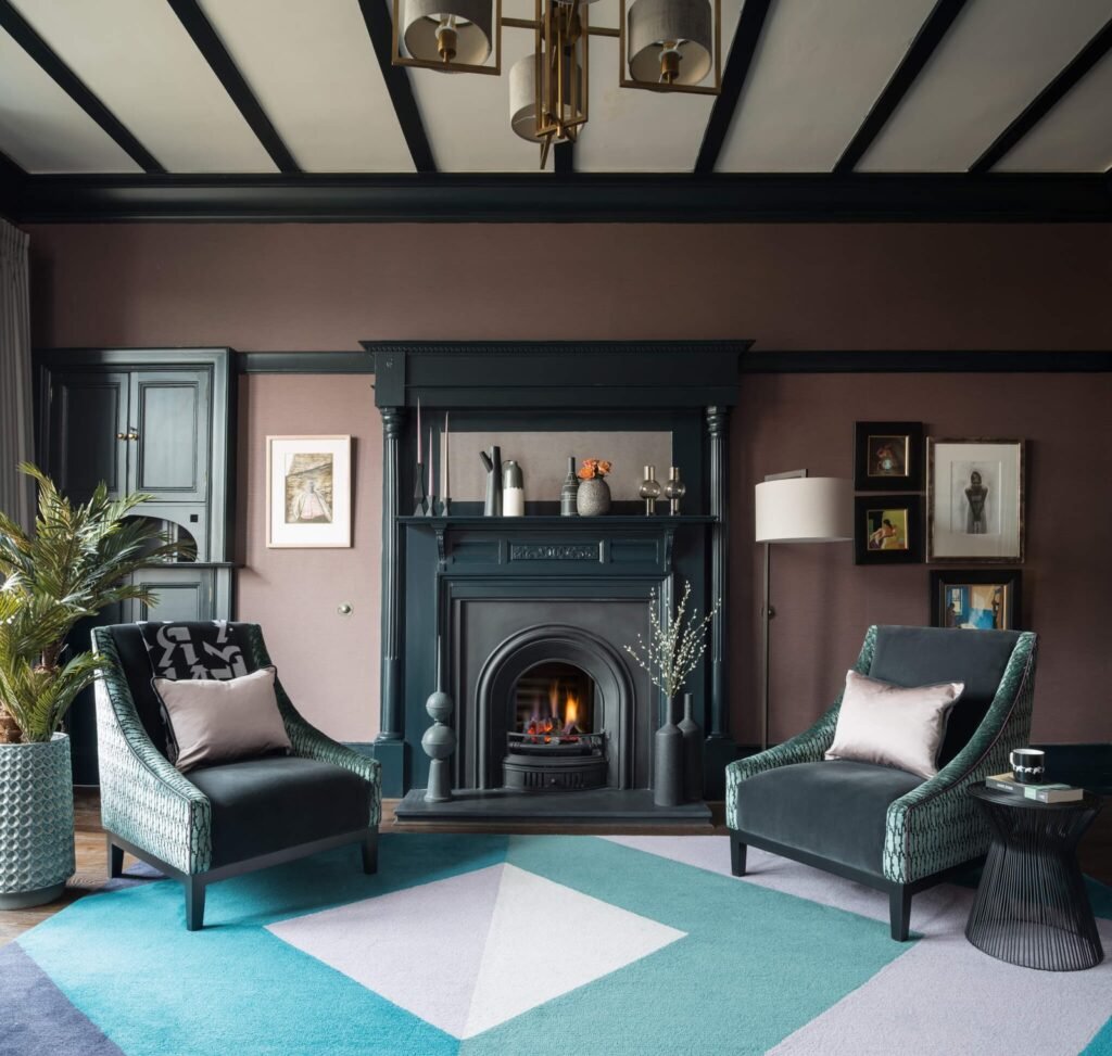 Elegant plum living room with a deep teal fireplace, two velvet armchairs, vibrant geometric rug, and beamed ceiling.