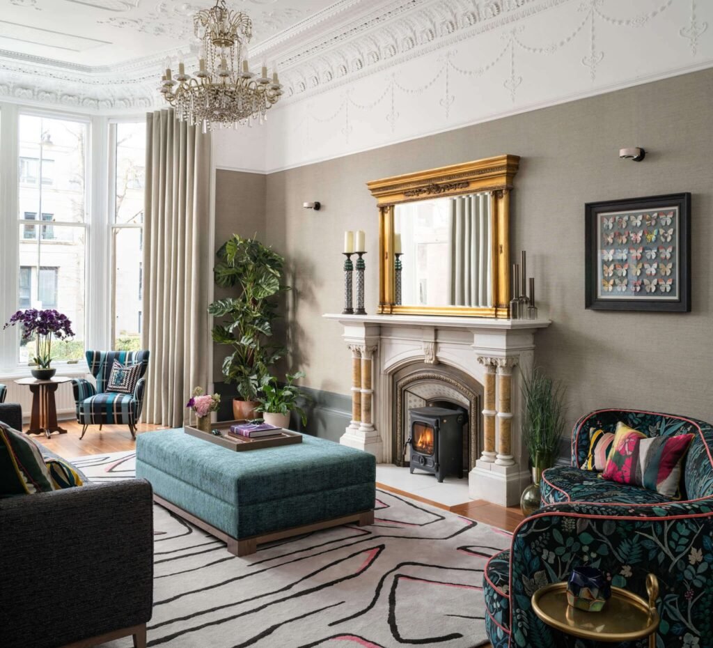 Eclectic living room with ornate fireplace, gold mirror, teal ottoman, patterned chairs, chandelier, and bay window.