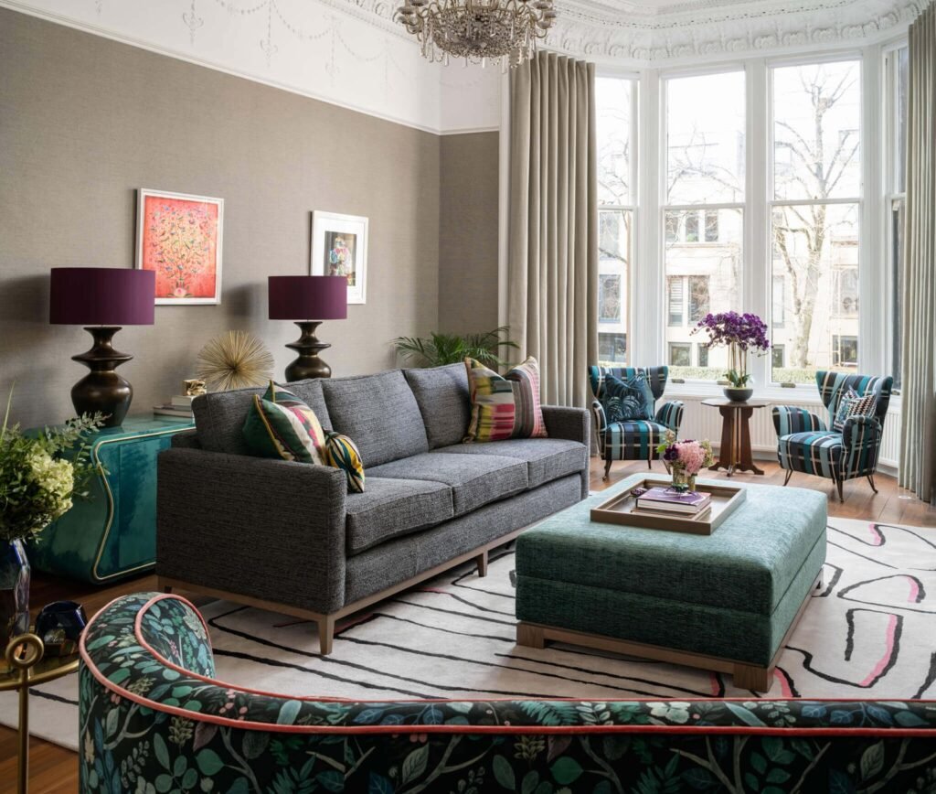 Stylish eclectic living room with grey sofa, plaid armchairs, green ottoman, dark floral bench, ornate ceiling & bay windows.