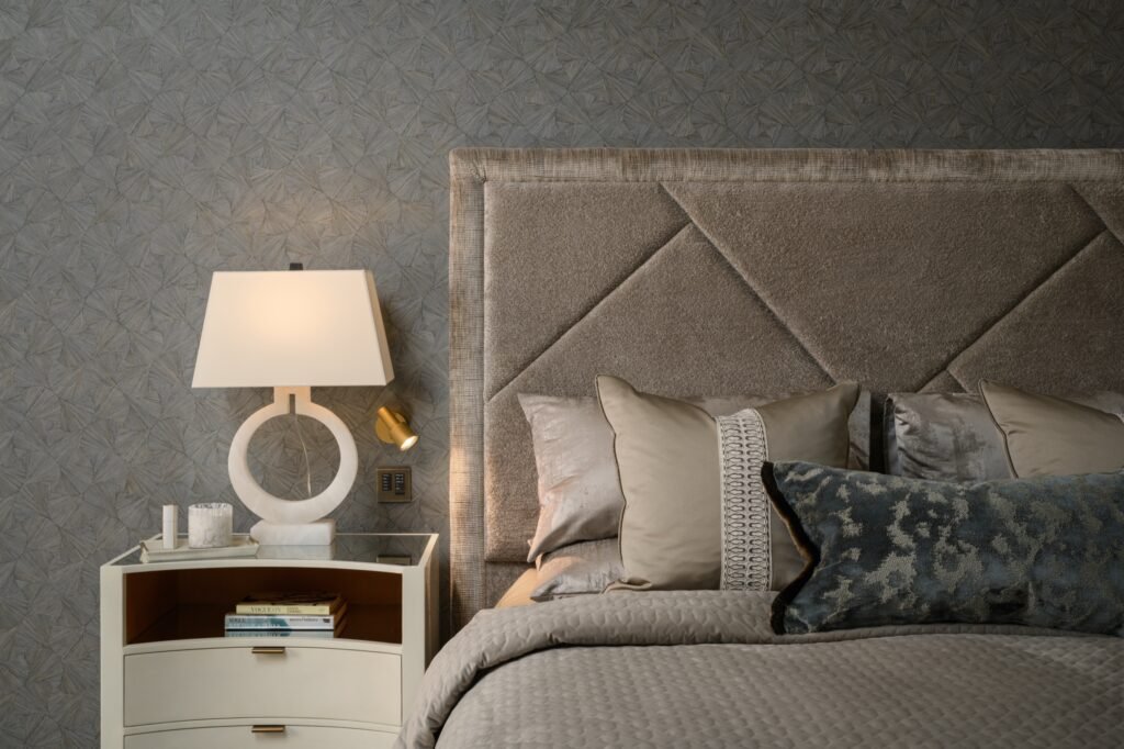 Elegant bedroom with textured geometric wallpaper, a geometric upholstered headboard, cream nightstand with lamp, and decorative pillows.