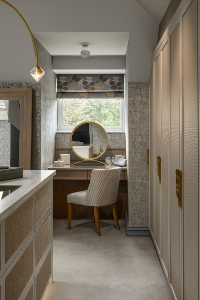Elegant dressing room with a wooden vanity, round gold mirror, patterned chair, floral window blind, textured walls, and custom wardrobes.