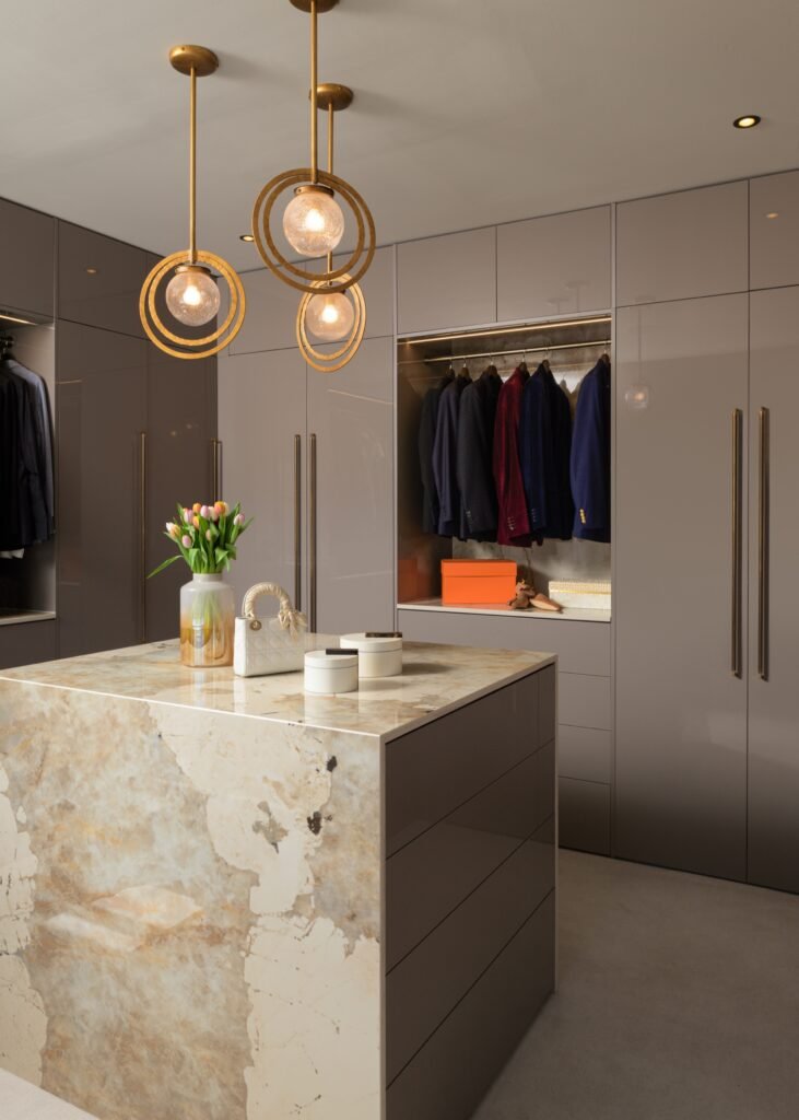 Modern luxury walk-in closet featuring taupe built-in wardrobes, a veined marble island, and unique gold pendant lights.