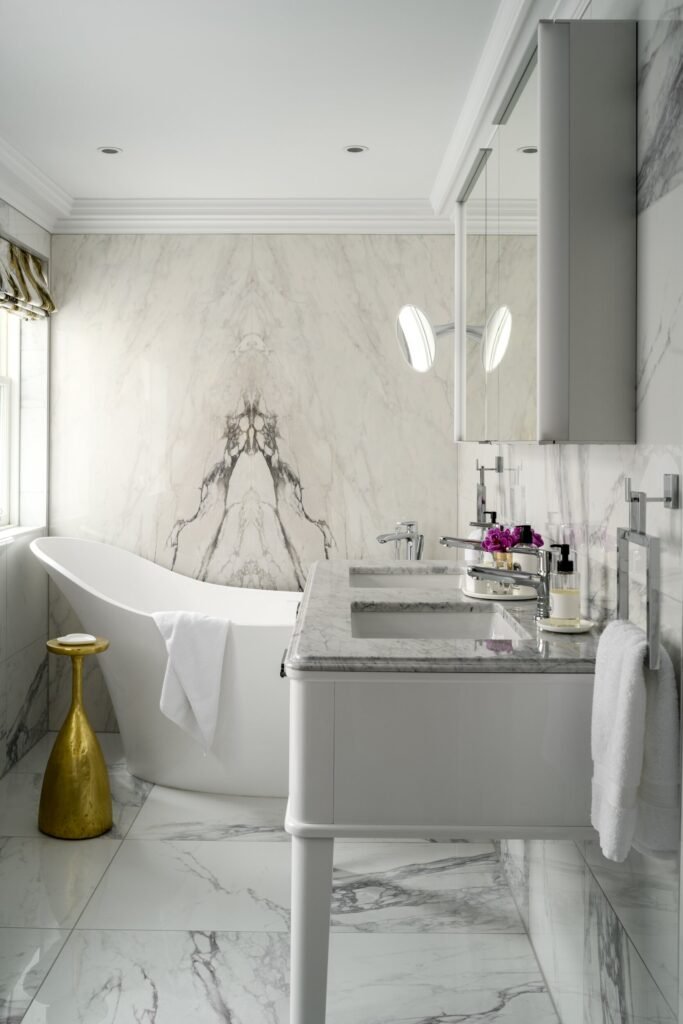 Luxurious white marble bathroom with a freestanding tub, double vanity, chrome fixtures, and striking bookmatched marble wall.