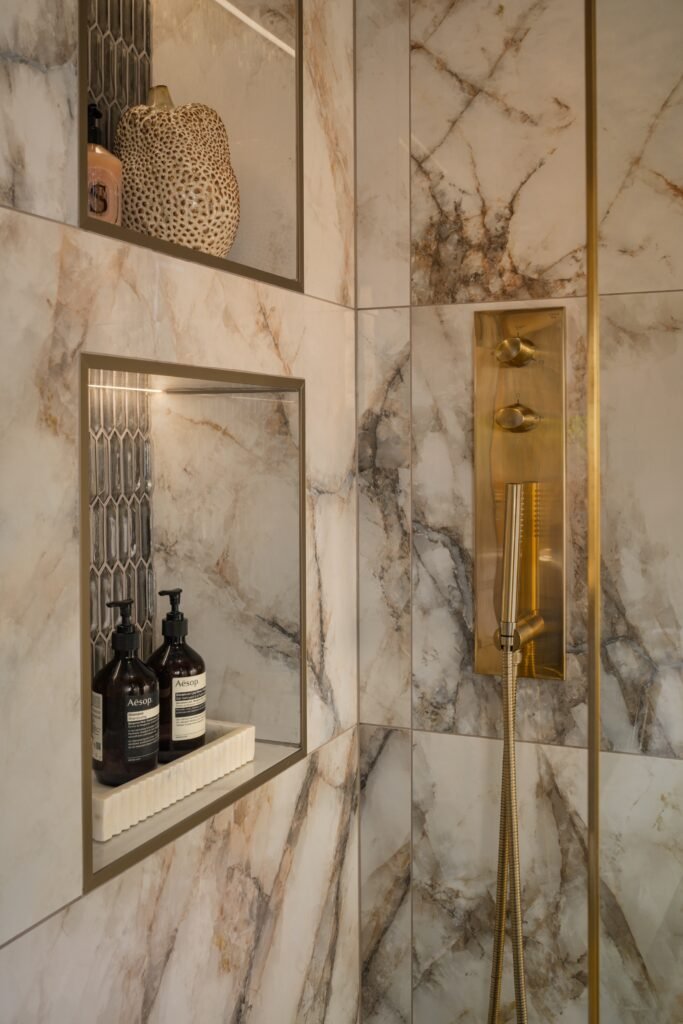 Modern shower with veined marble tiles, brass shower controls, and illuminated niches holding Aesop products and decor.