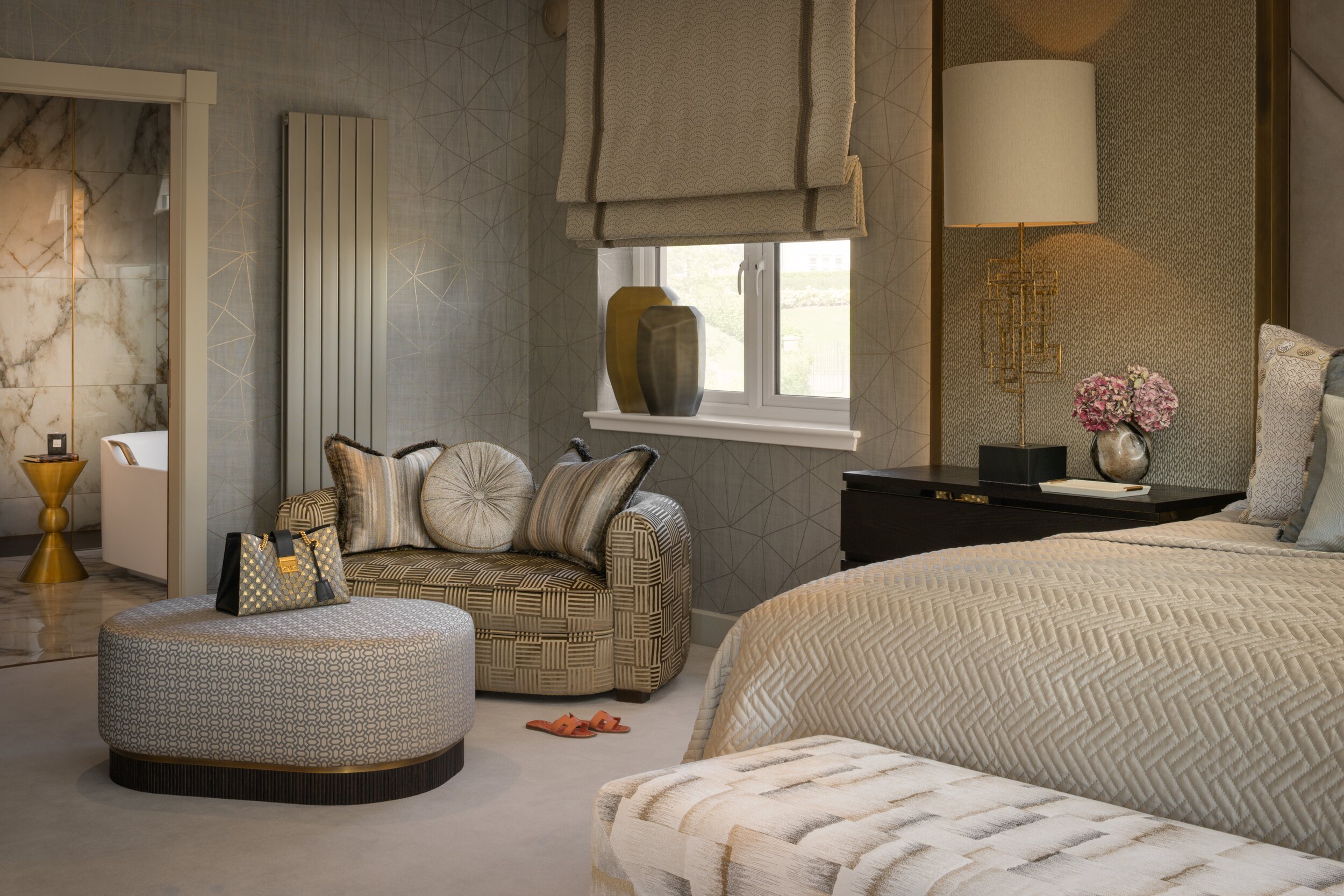 Sophisticated bedroom with geometric gray and gold wallpaper, patterned seating, large bed, and a marble en-suite bathroom.