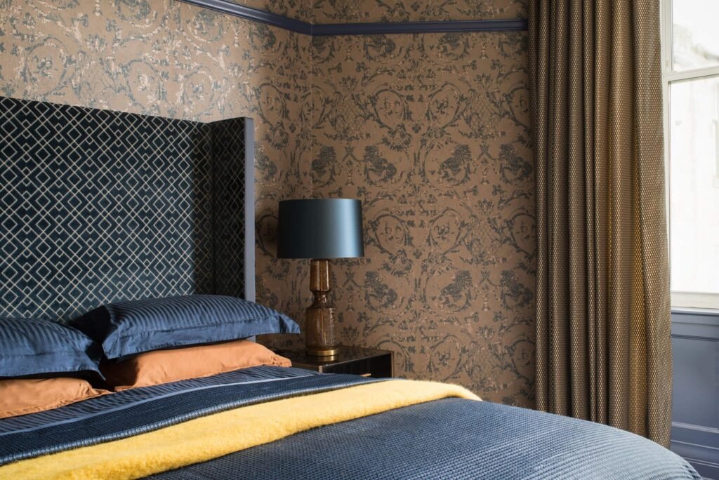 Elegant bedroom: blue patterned headboard, ornate brown & blue wallpaper, blue bedding, yellow throw, and bedside lamp.