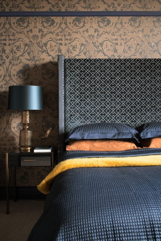 Luxurious bedroom with dark blue geometric velvet headboard, ornate taupe wallpaper, gold lamp, and colorful bedding.