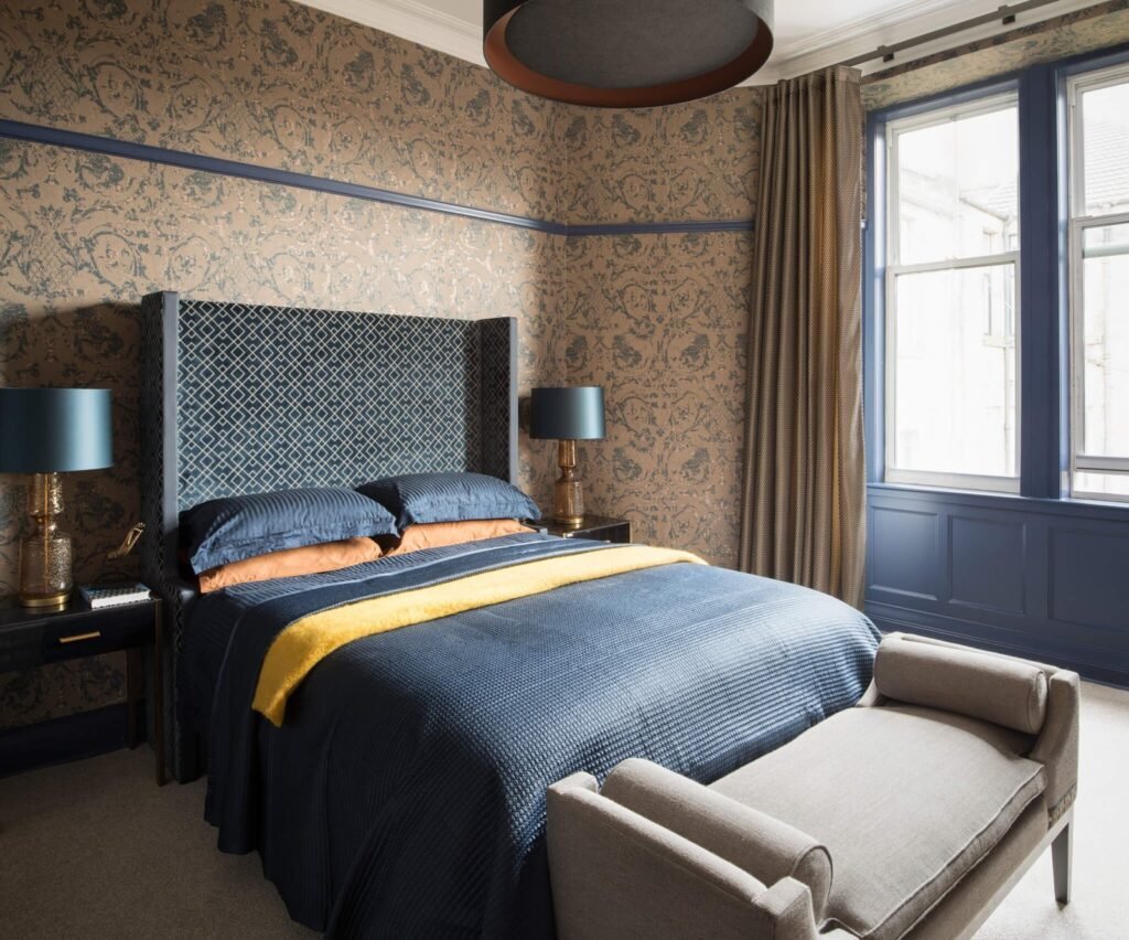 Elegant bedroom with navy blue quilted bed, patterned headboard, ornate beige wallpaper, and dark blue window trim.
